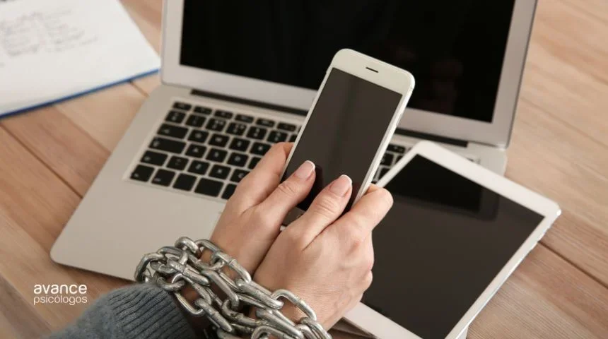 Manos encadenadas sujetando un móvil frente a una tablet y un portátil, representando la adicción a la tecnología y redes sociales.